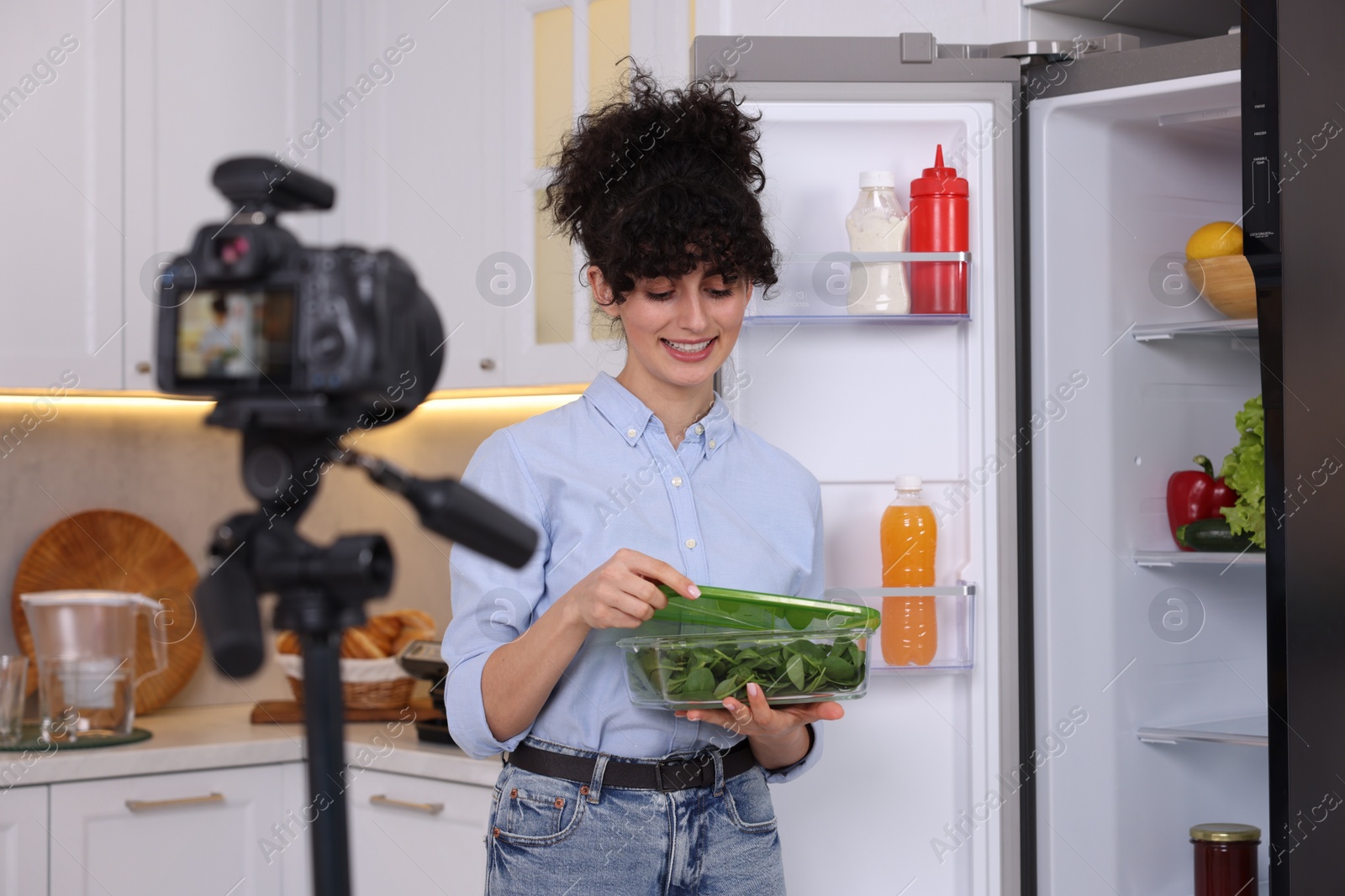 Photo of Smiling food blogger opening container with spinach while recording video in kitchen