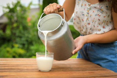 Woman pouring fresh milk from can into glass at wooden table outdoors, closeup