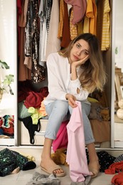 Photo of Young woman sitting in wardrobe with different clothes indoors. Fast fashion concept