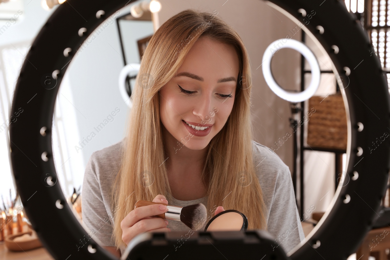 Photo of Beautiful young woman applying face powder with brush indoors, view through ring lamp