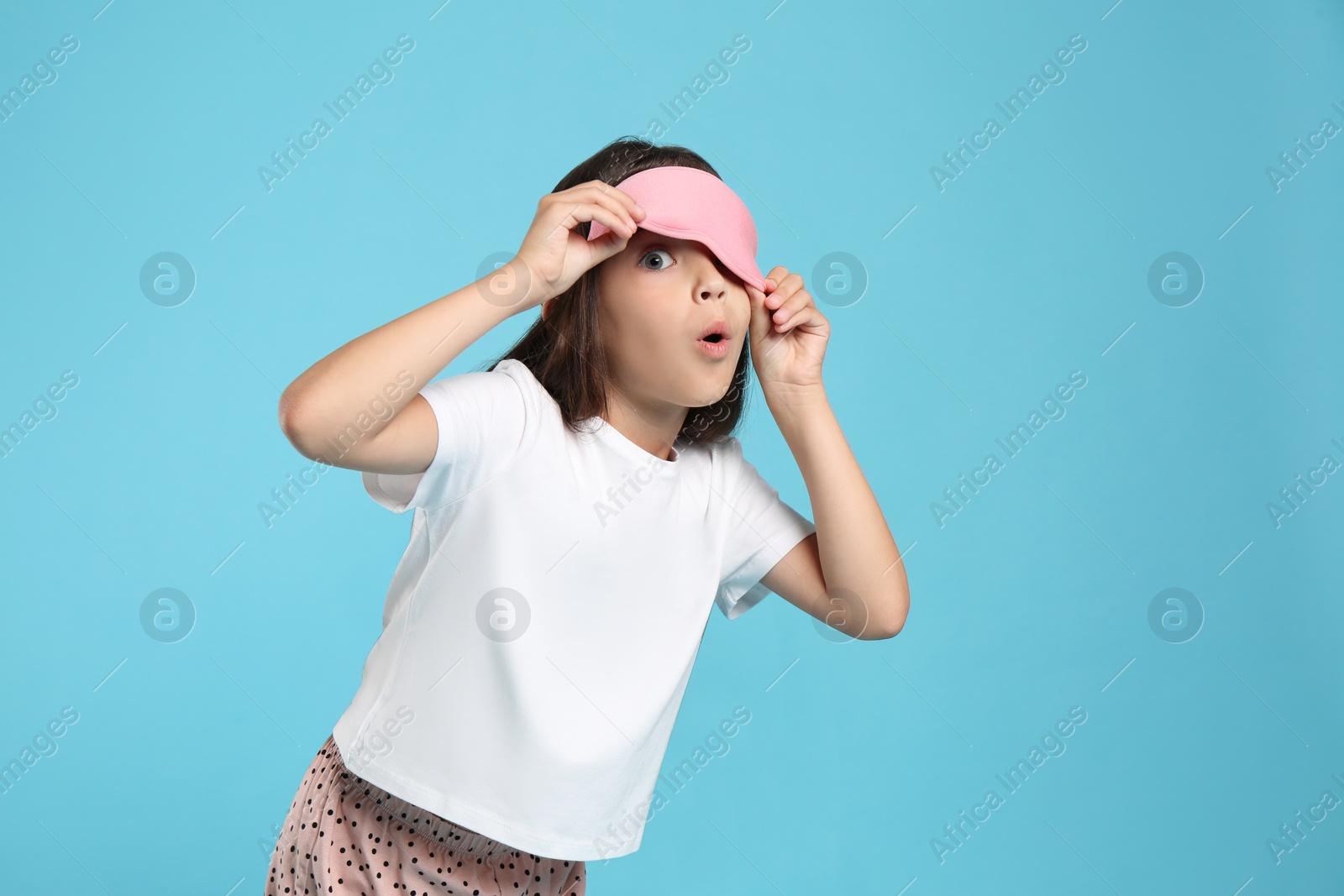 Photo of Cute girl wearing pajamas and sleeping mask on light blue background