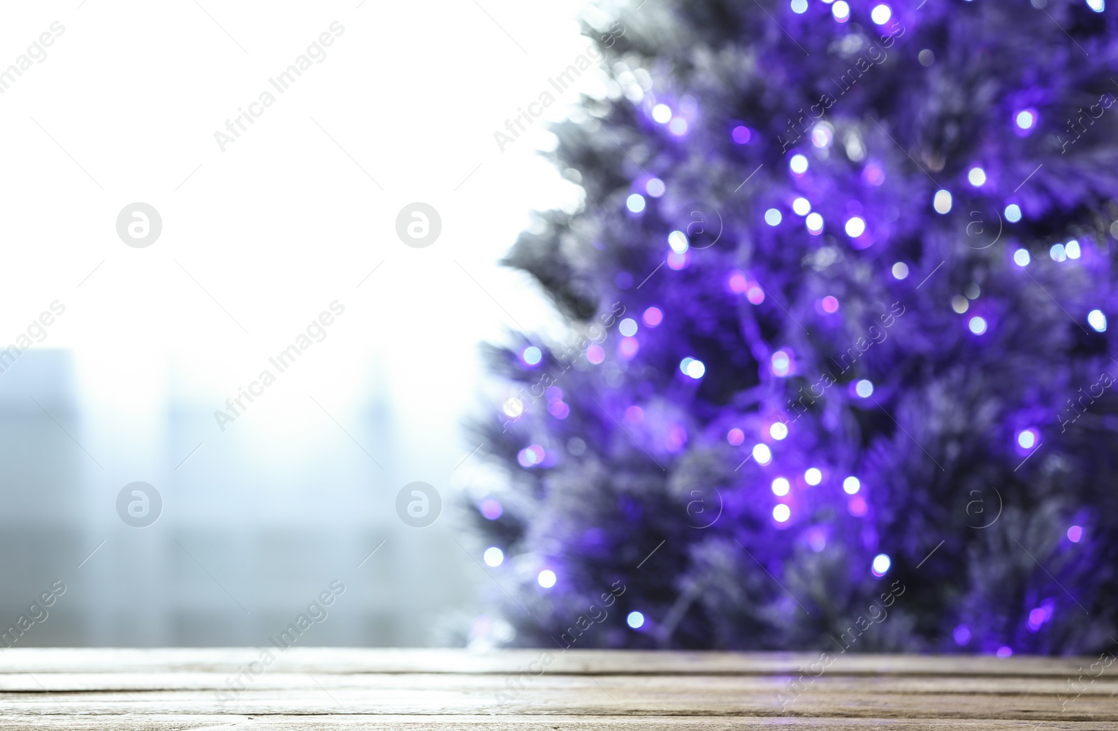 Photo of Blurred view of beautiful Christmas tree with purple lights near window indoors, focus on wooden table. Space for text