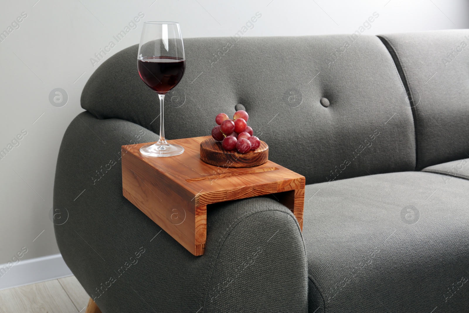 Photo of Glass of red wine and grapes on sofa with wooden armrest table in room. Interior element