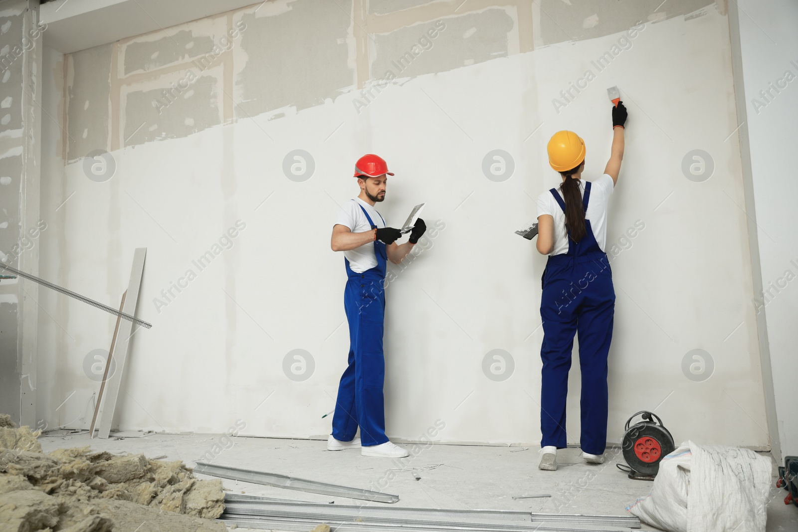 Photo of Professional workers plastering wall with putty knives in hard hats indoors
