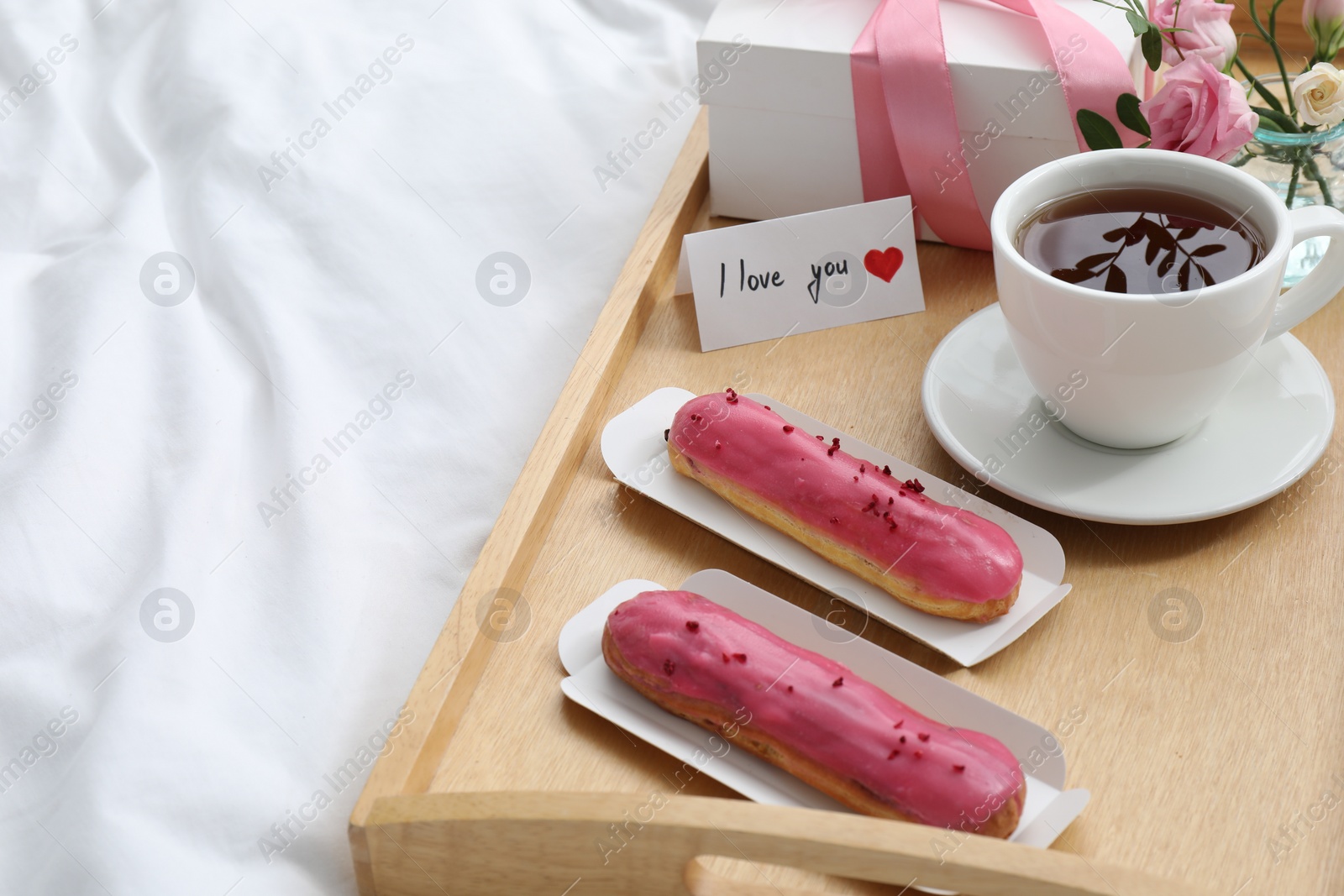 Photo of Tasty breakfast served in bed. Delicious eclairs, tea, gift box, flowers and card with phrase I Love You on tray