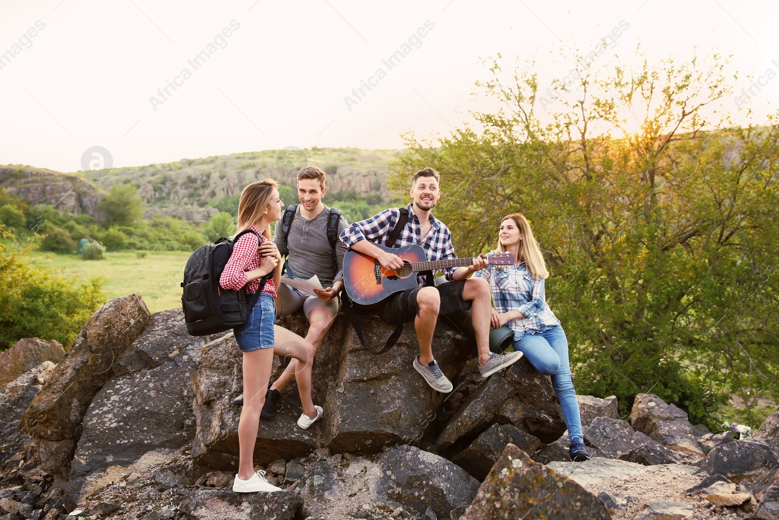 Photo of Young man with backpack playing guitar for his friends in wilderness. Camping season