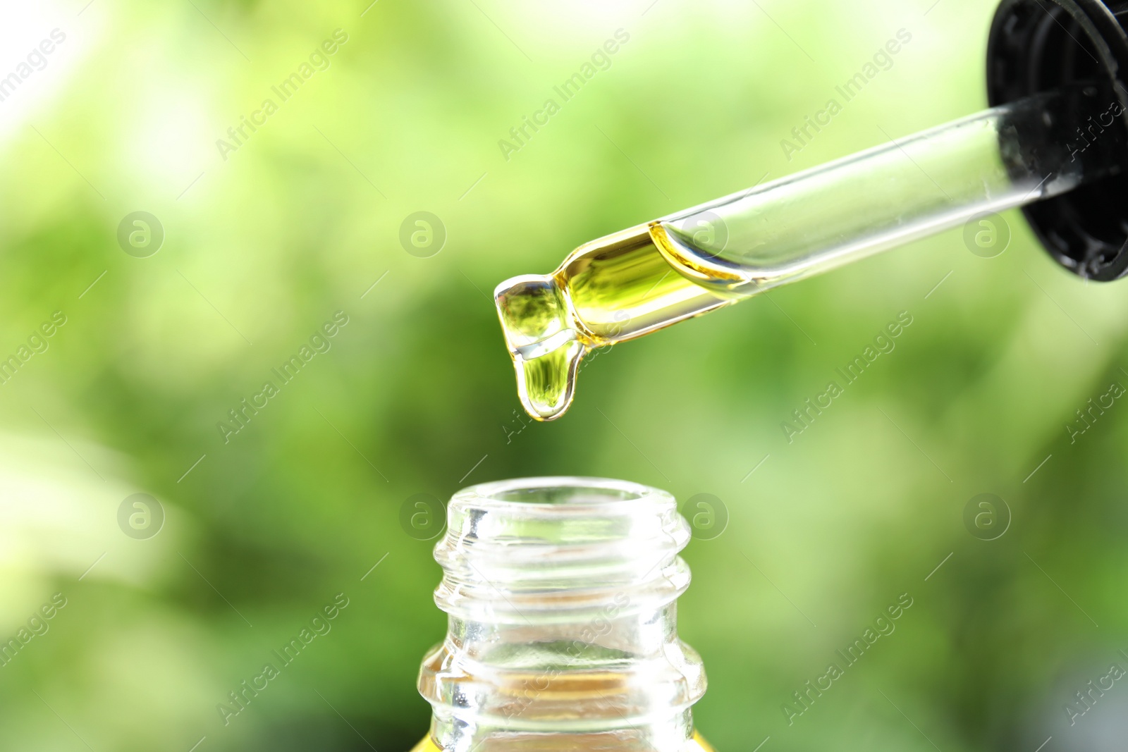 Photo of Dripping essential oil from pipette into glass bottle against blurred green background, closeup