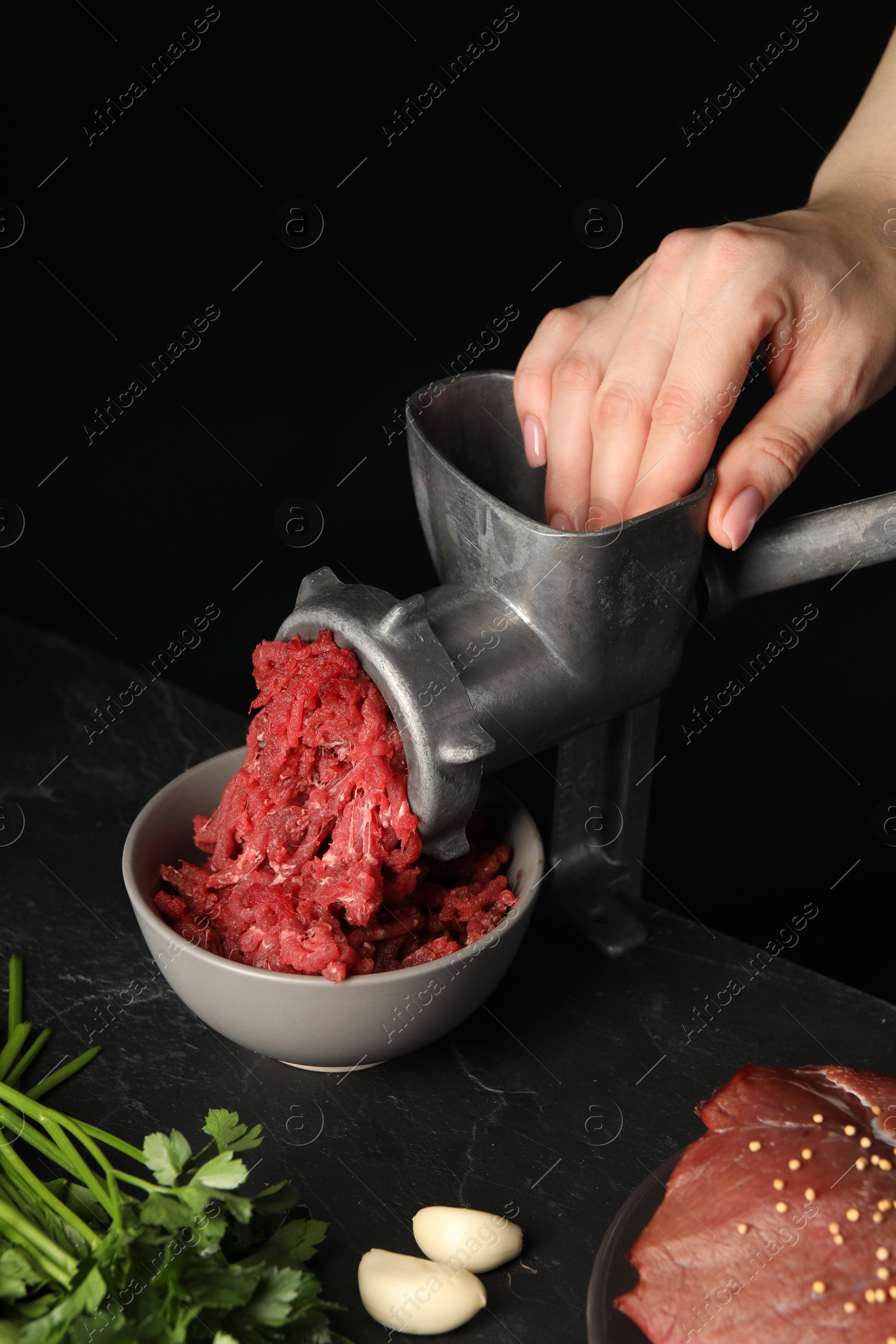 Photo of Woman making beef mince with manual meat grinder at dark textured table against black background, closeup