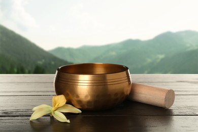 Image of Golden singing bowl, mallet and flower on wooden table against mountain landscape