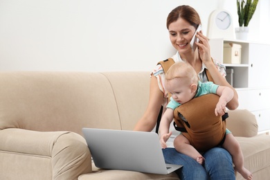 Photo of Woman with her son in baby carrier using laptop and talking on phone at home. Space for text