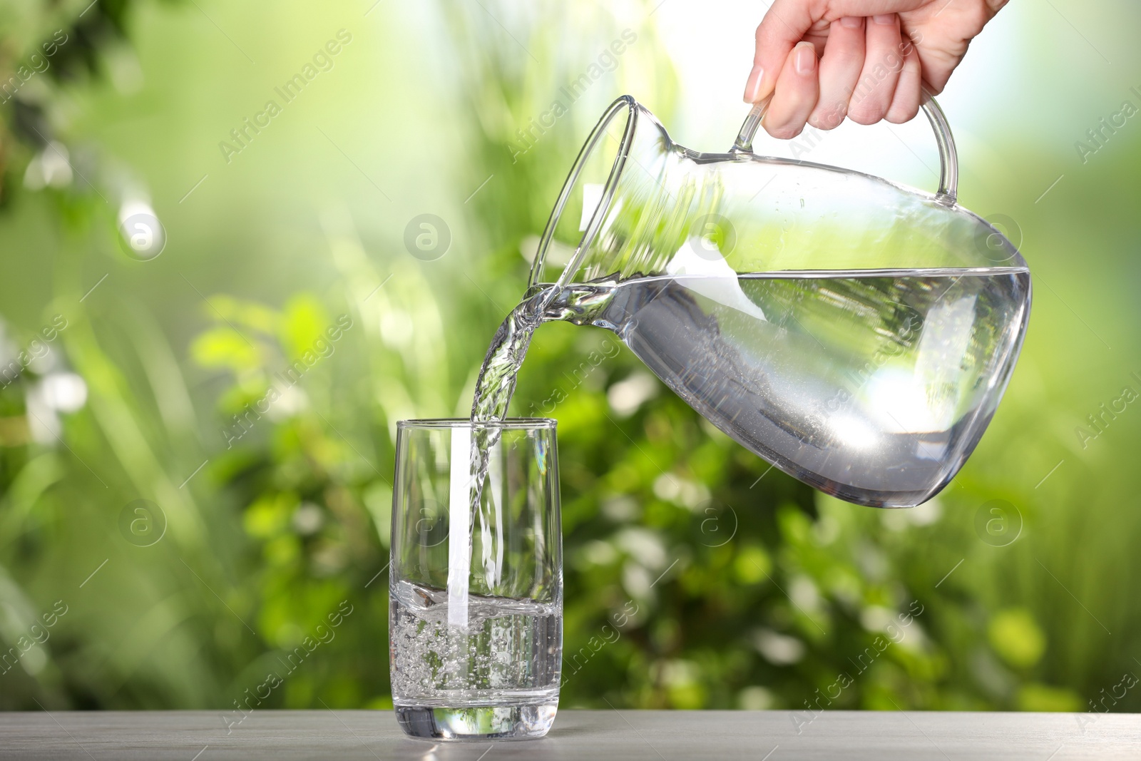 Photo of Woman pouring water from jug into glass on light grey table outdoors, closeup