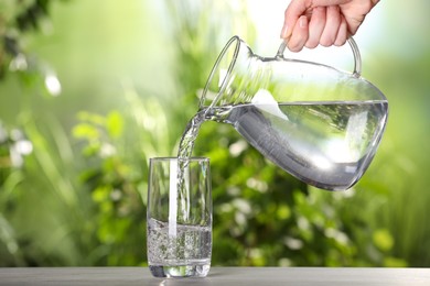 Photo of Woman pouring water from jug into glass on light grey table outdoors, closeup