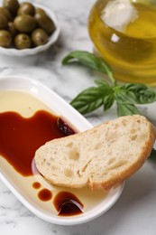 Photo of Bowl of organic balsamic vinegar with oil served with bread slice, basil and olives on white marble table