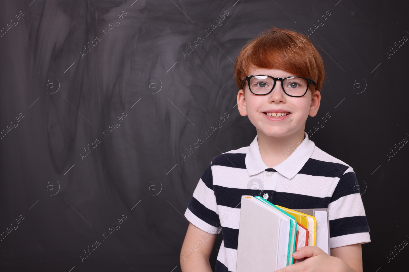 Photo of Smiling schoolboy in glasses with books near blackboard. Space for text