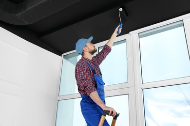 Worker in uniform painting ceiling with roller on stepladder indoors