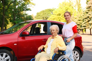 Photo of Senior woman in wheelchair with granddaughter near car outdoors