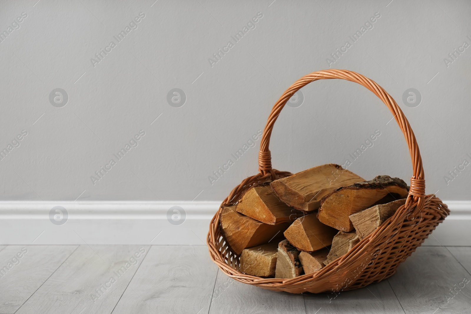 Photo of Wicker basket with firewood near grey wall indoors, space for text