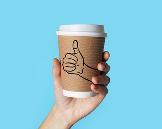 Image of Woman holding takeaway paper cup on light blue background, closeup. Thumbs up gesture drawing on cup