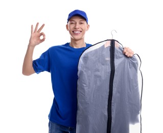 Photo of Dry-cleaning delivery. Happy courier holding garment cover with clothes and showing OK gesture on white background
