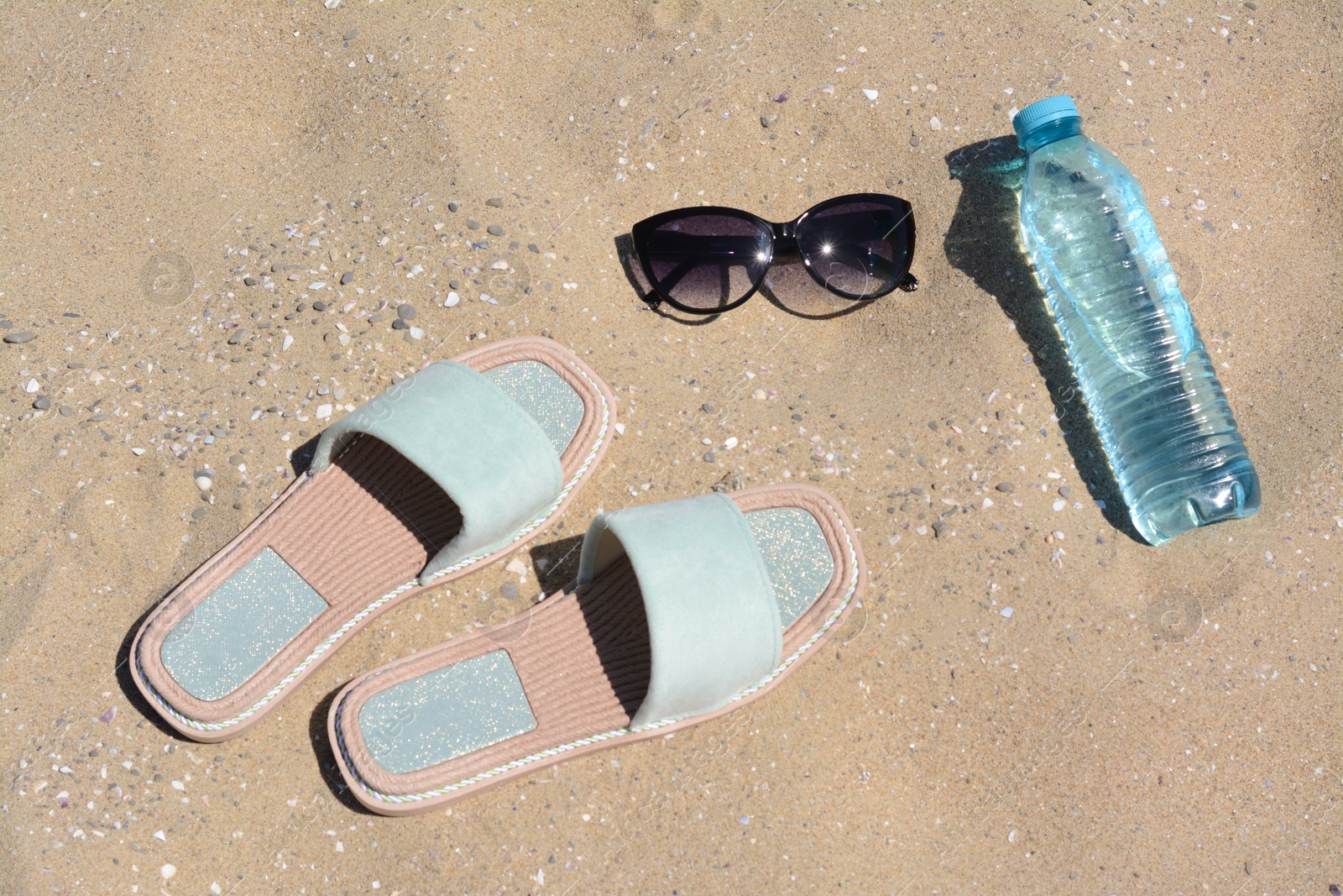 Photo of Stylish sunglasses, slippers and bottle of water on sand. Beach accessories