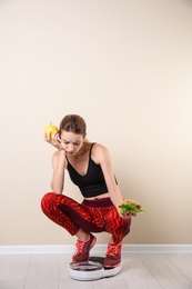 Photo of Woman holding tasty sandwich and apple while measuring her weight on floor scales near light wall. Weight loss motivation