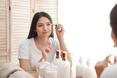 Beautiful woman applying oil onto her eyelashes near mirror indoors