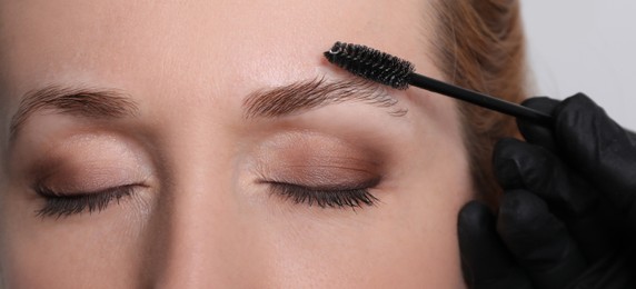 Photo of Beautician brushing woman's eyebrows before tinting on light grey background, closeup