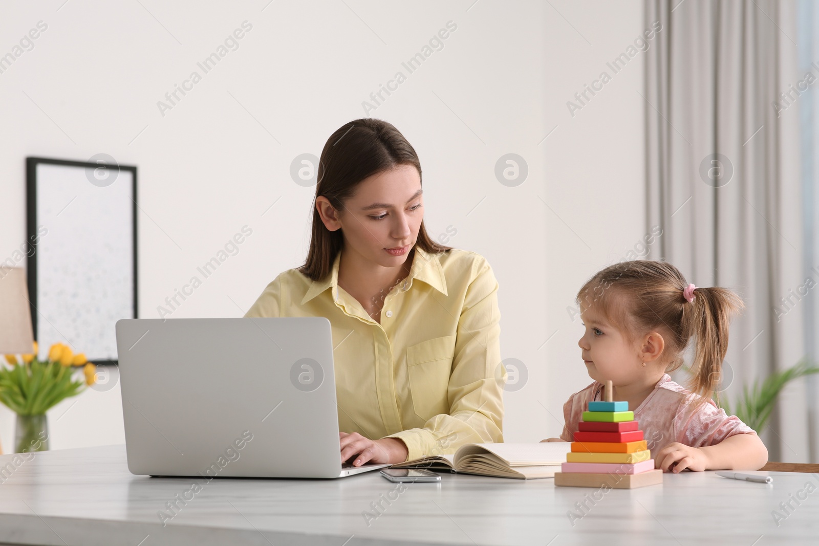 Photo of Woman working remotely. Mother with laptop and daughter at desk