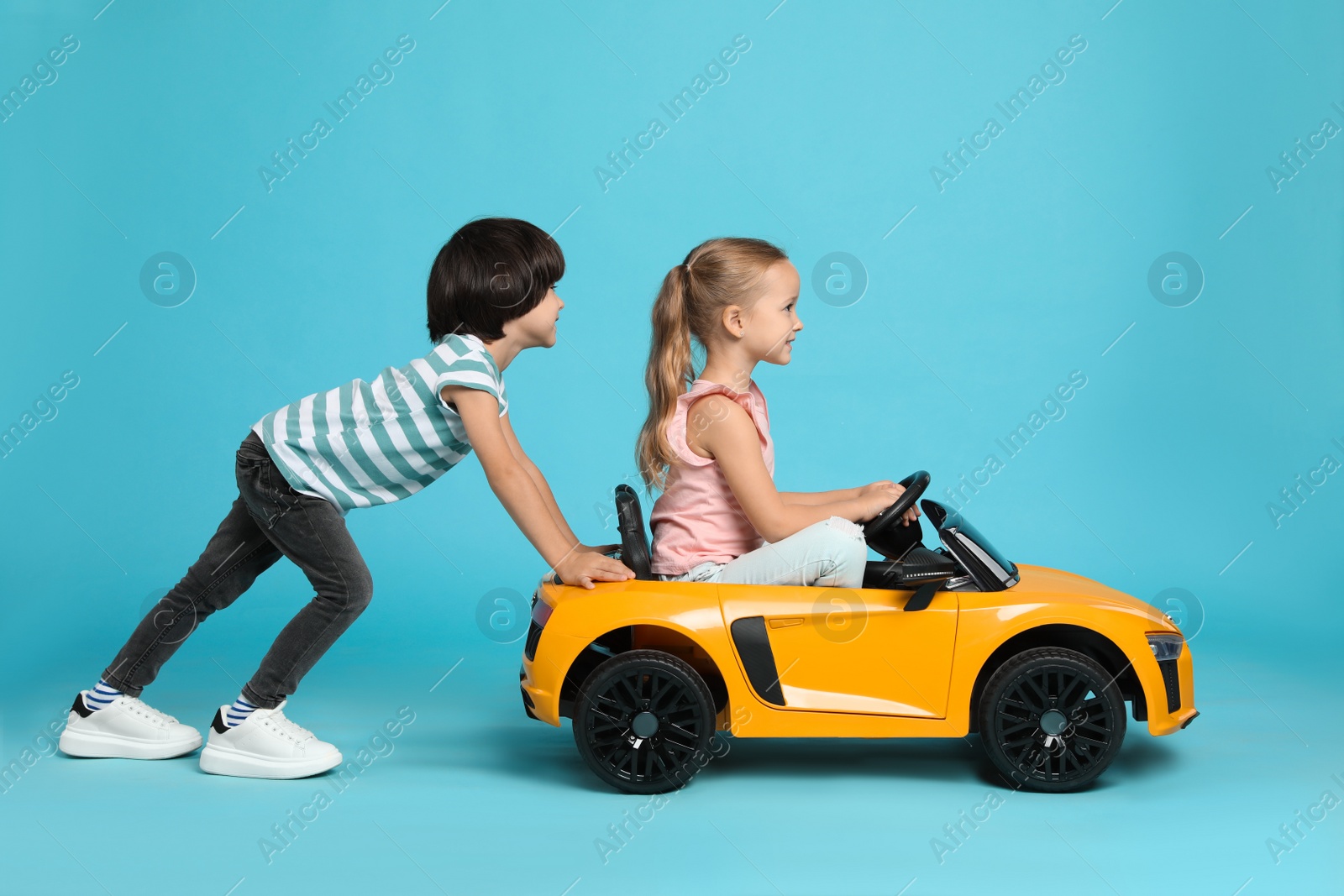 Photo of Cute boy pushing children's electric toy car with little girl on light blue background