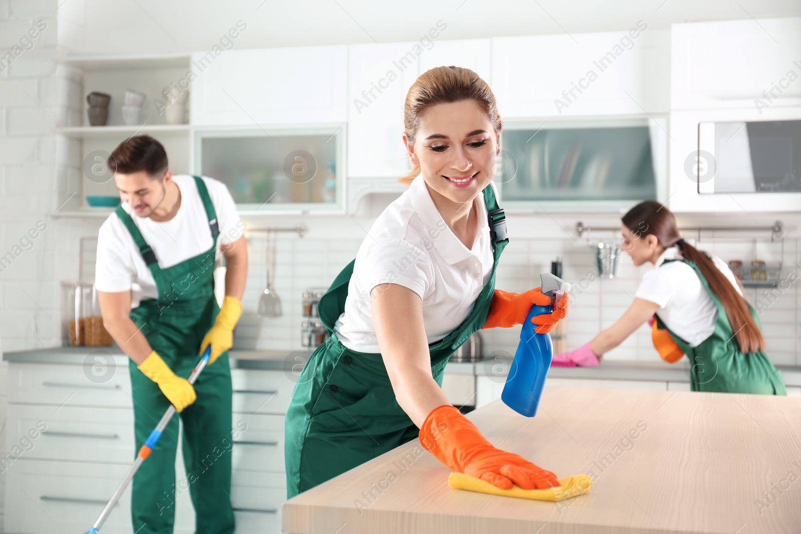 Photo of Woman using rag and sprayer for cleaning table with colleagues in kitchen