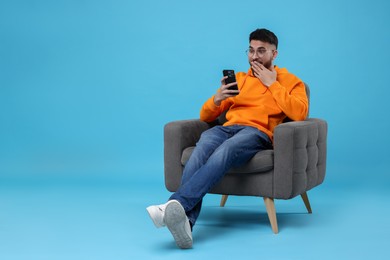 Emotional young man using smartphone on armchair against light blue background, space for text