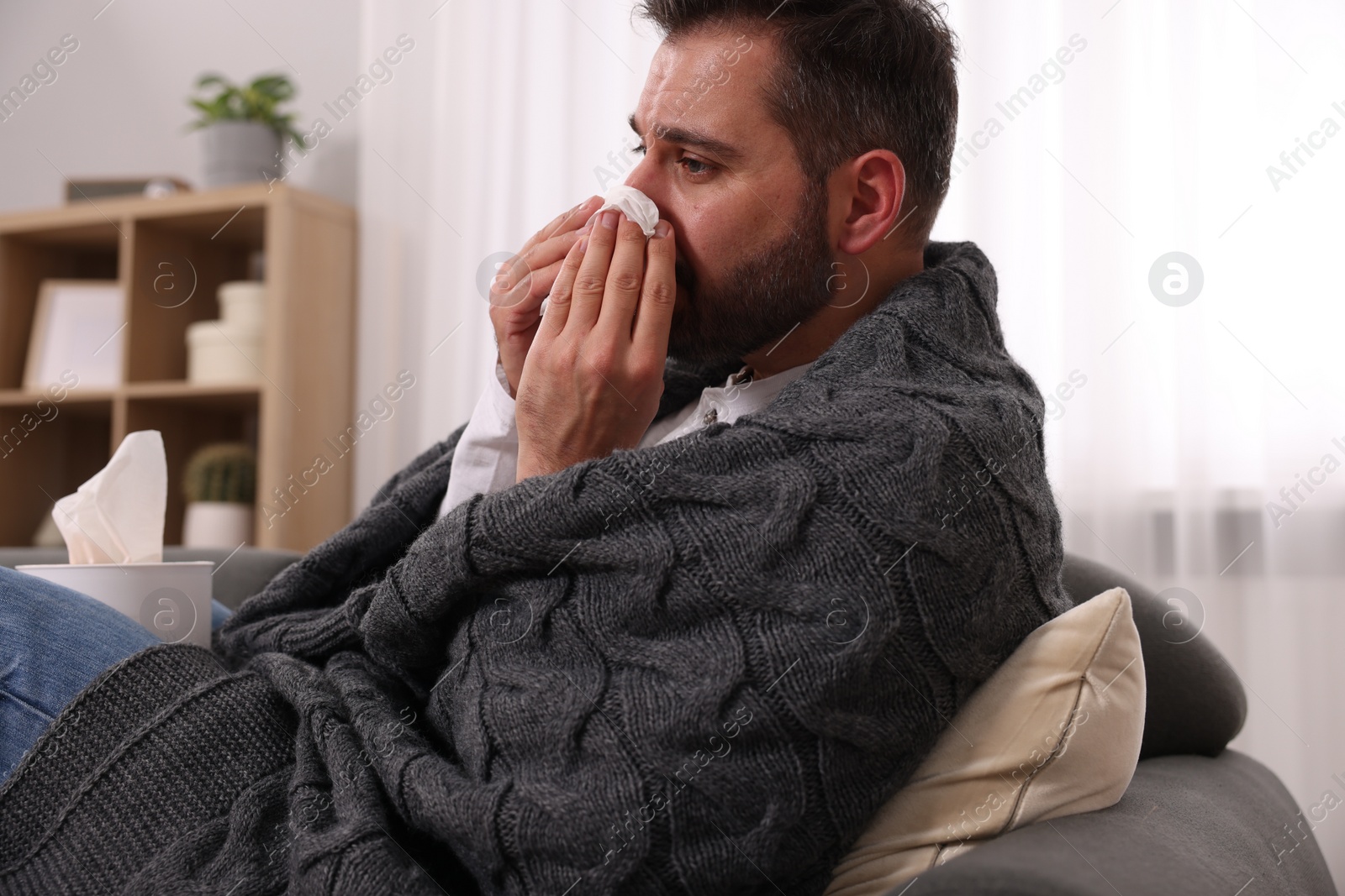 Photo of Sick man wrapped in blanket with tissue blowing nose on sofa at home. Cold symptoms