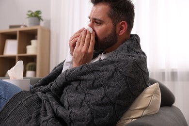 Photo of Sick man wrapped in blanket with tissue blowing nose on sofa at home. Cold symptoms