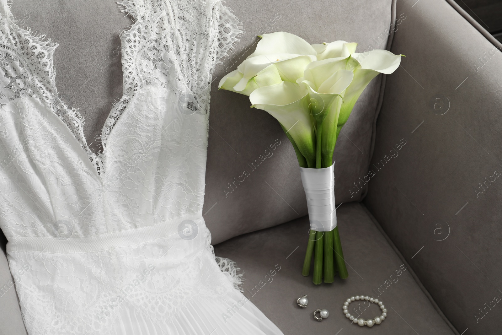 Photo of Beautiful calla lily flowers tied with ribbon, wedding dress and jewelry on sofa