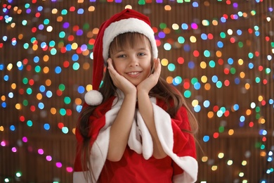 Photo of Cute little child in Santa hat on blurred lights background. Christmas celebration