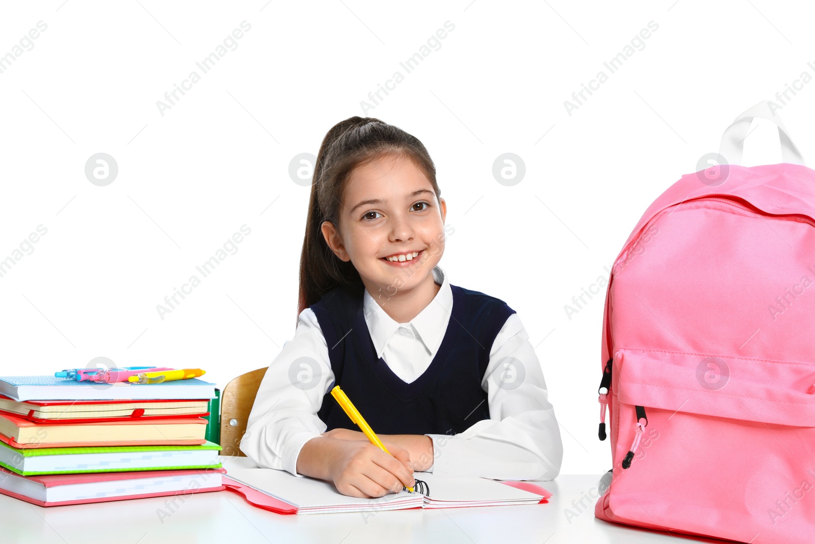 Photo of Little girl in uniform doing assignment at desk against white background. School stationery