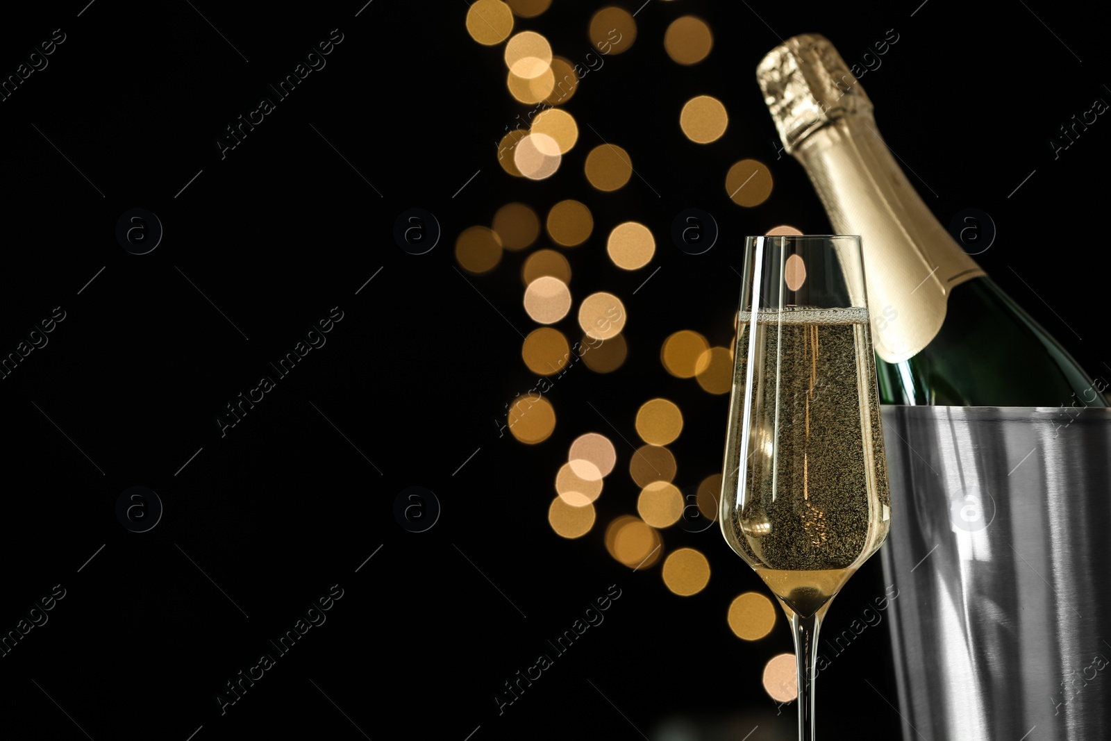 Photo of Glass of champagne near bucket with bottle against blurred lights, space for text