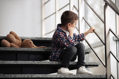 Photo of Sad little boy with toy sitting on stairs indoors