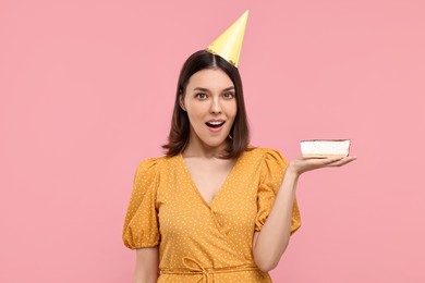 Surprised woman in party hat with cheesecake on pink background