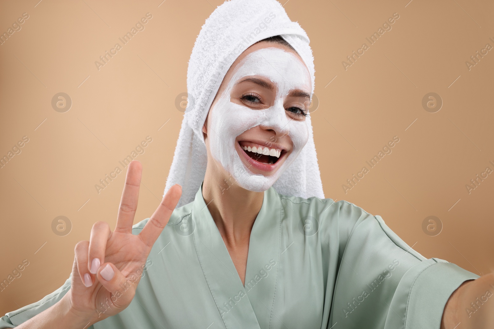 Photo of Woman with face mask showing v-sign while taking selfie on beige background. Spa treatments