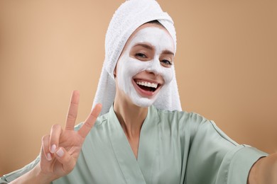 Photo of Woman with face mask showing v-sign while taking selfie on beige background. Spa treatments