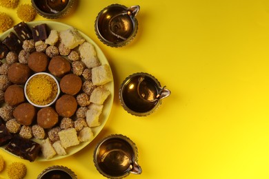 Happy Diwali. Flat lay composition with diya lamps, chrysanthemum flowers and delicious Indian sweets on yellow table. Space for text