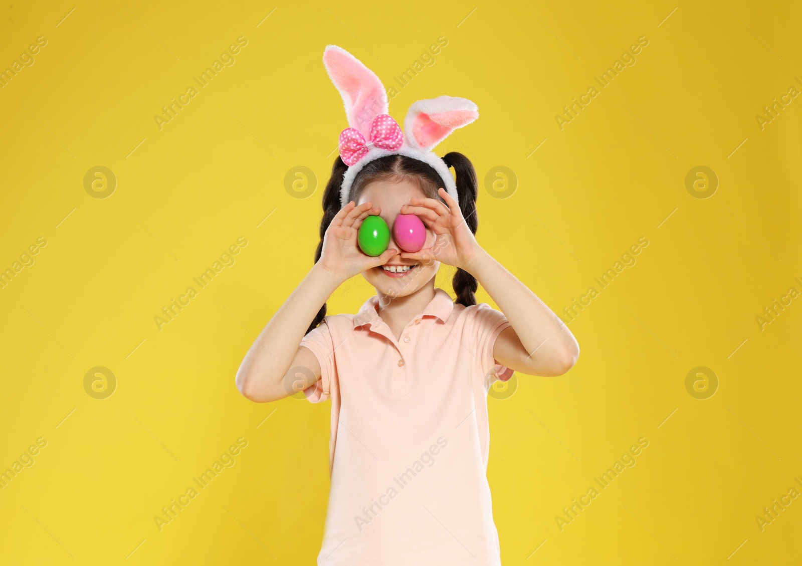 Photo of Little girl in bunny ears headband holding Easter eggs near eyes on color background
