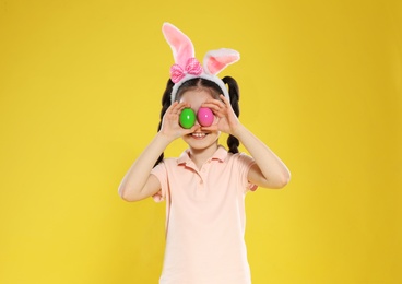 Little girl in bunny ears headband holding Easter eggs near eyes on color background