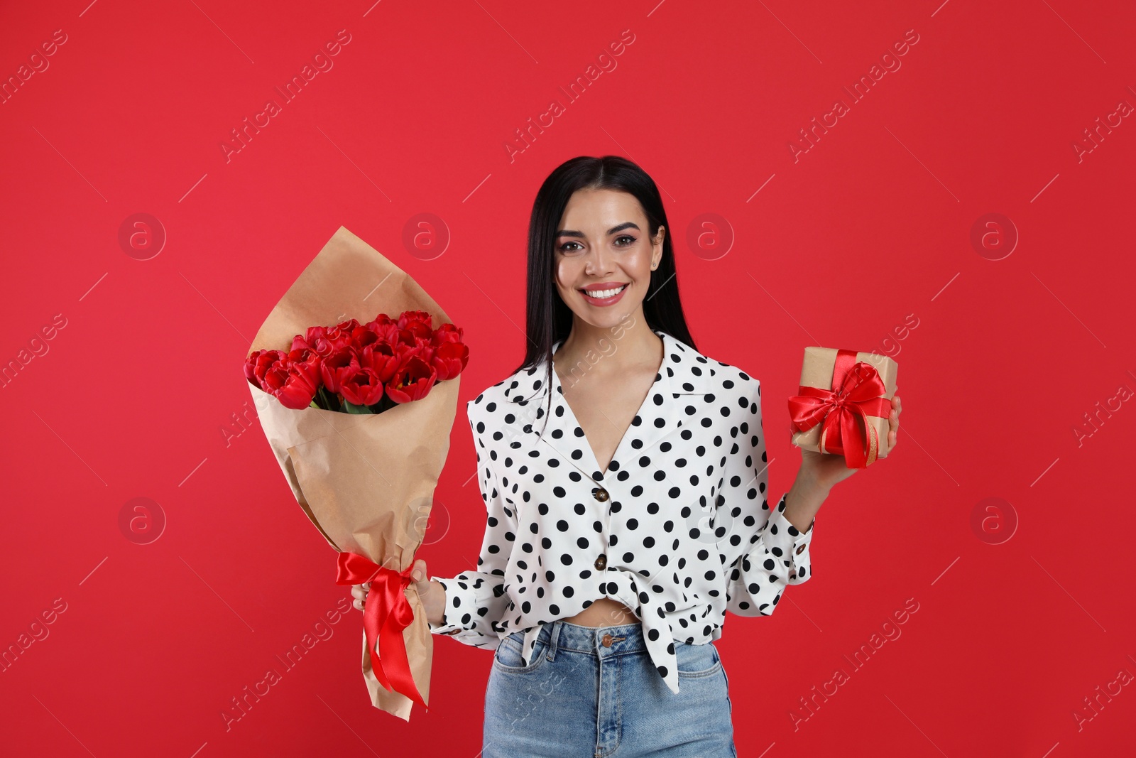 Photo of Happy woman with tulip bouquet and gift box on red background. 8th of March celebration
