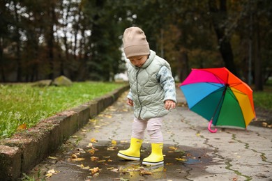 Photo of Cute little girl walking in puddle near colorful umbrella outdoors