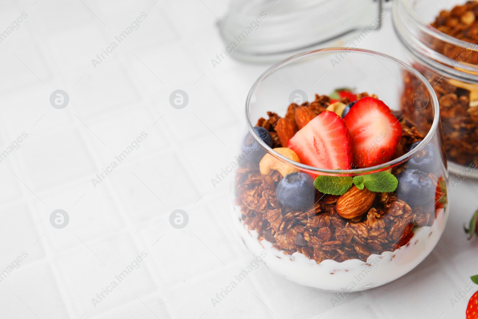 Photo of Tasty granola with berries, nuts and yogurt in glass on white tiled table, closeup. Space for text