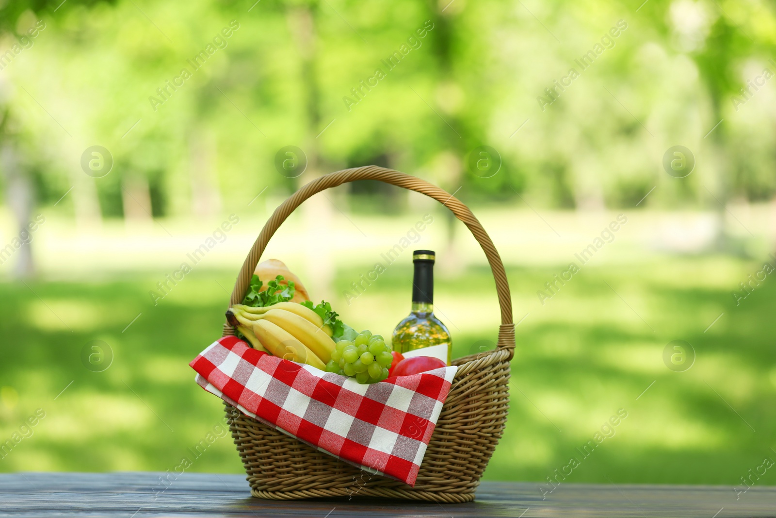 Photo of Wicker basket with blanket, wine and food on table in park. Summer picnic