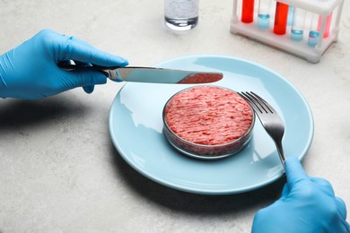 Photo of Scientist holding fork and knife over plate with minced cultured meat at light grey lab table, closeup