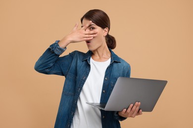 Embarrassed woman with laptop covering face on beige background
